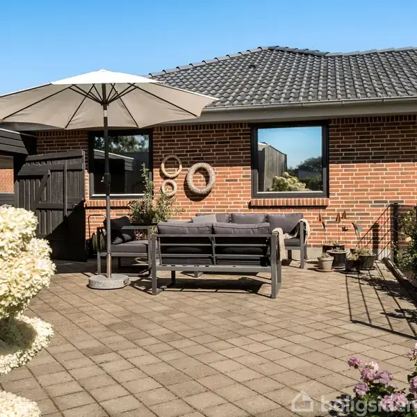 En terrasse med grå fliser har en grå sofa og parasol. En rød murstensbygning med vinduer danner baggrunden, omkranset af blomsterbede med lyserøde og hvide blomster.