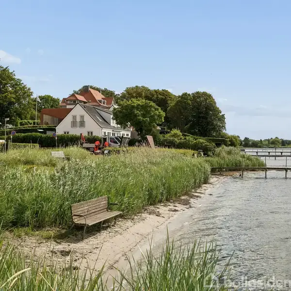 Bænk står stille ved kysten omgivet af højt græs og små huse. En bro strækker sig ud over vandet i en fredelig landskabsscene.