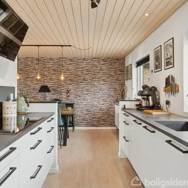 Et moderne køkken med hvide skabe og sorte håndtag. Der står en kaffemaskine ved vinduet, og en spiseplads ses i baggrunden med lamper hængende over et bord.