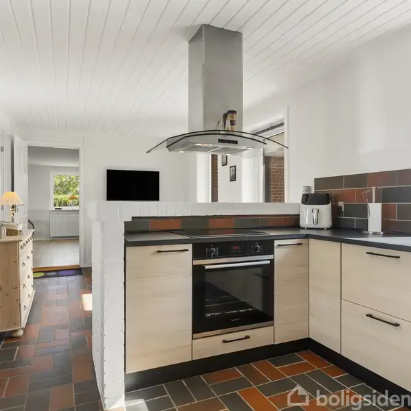 Et moderne køkken med lyse træskabe og sorte bordplader. En indbygget ovn og emhætte står i midten. Køkkenet er omgivet af vinduer, der giver naturligt lys. Mørkt flisegulv.