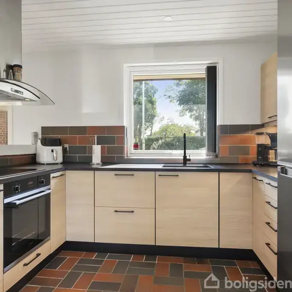 Køkken med lyse træskabe og sorte bordplader. En sort håndvask er under et vindue, der viser træer udenfor. Et stålkomfur og køleskab står til højre. Brug af farvestrålende fliser på gulv og væg.