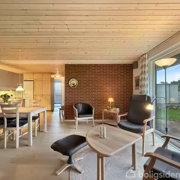 Stue med træmøbler, hvor stole er arrangeret omkring et rundt sofabord. Indretningen domineres af en murstensvæg. Store vinduer giver udsigt til en have.