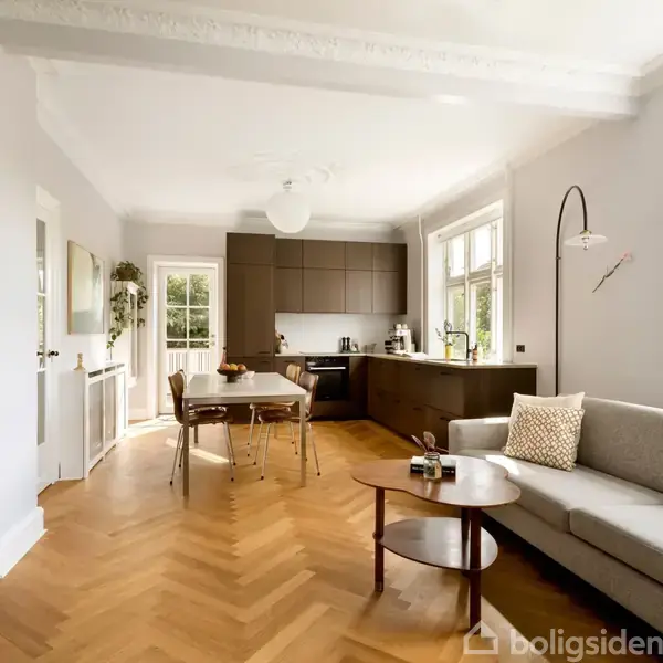Stue med en grå sofa og et rundt træbord. Køkken i baggrunden med moderne skabsdesign. Spisebord og stole ved siden af sofaen. Rummet har lyse vægge og trægulv.
