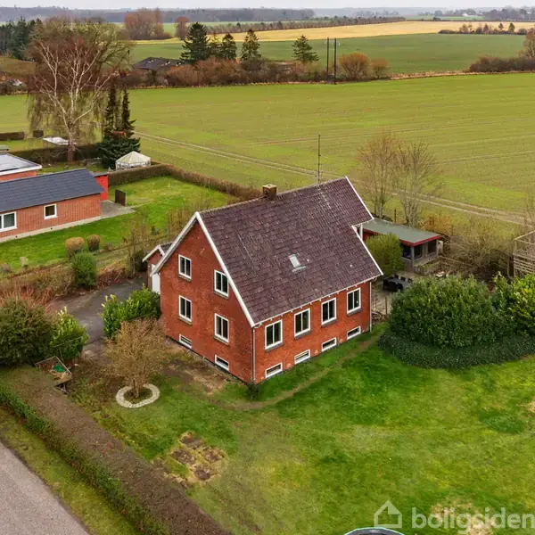 En stue med en blå sofa står i et hjørne ved siden af et træbord. I baggrunden ses en spiseplads med stole og et brændekomfur omgivet af et sikkerhedsgelænder.