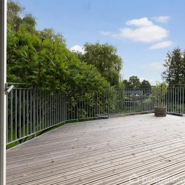 En åben terrasse med trægulv omgivet af et metalgelænder. I baggrunden er der grønne træer og buske under en skyet, blå himmel.