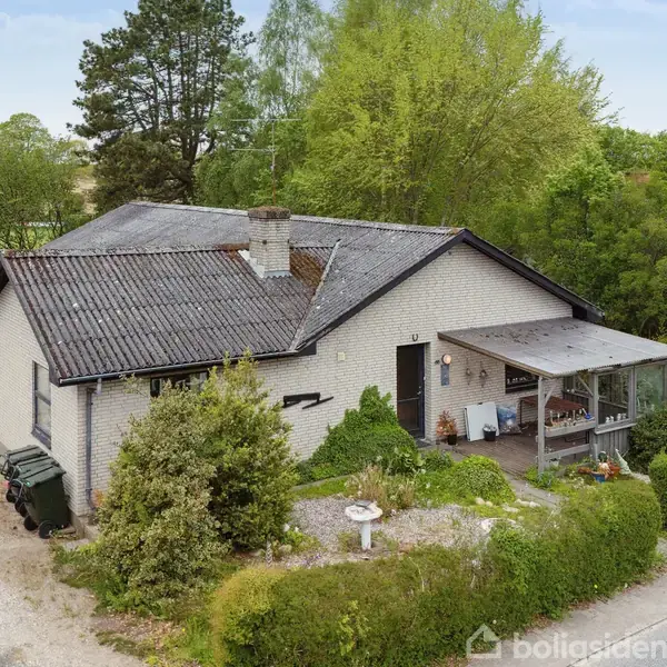 Hus med gråt tag og murede vægge, har en overdækket veranda med møbler. Beliggende i en grøn, frodig have med buske og grusvej foran.