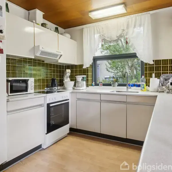 Et lyst, funktionelt køkken med hvide skabe og grønne flisevægge. En ovn og mikrobølgeovn står ved siden af hinanden. Store vinduer i baggrunden giver udsigt til en have.