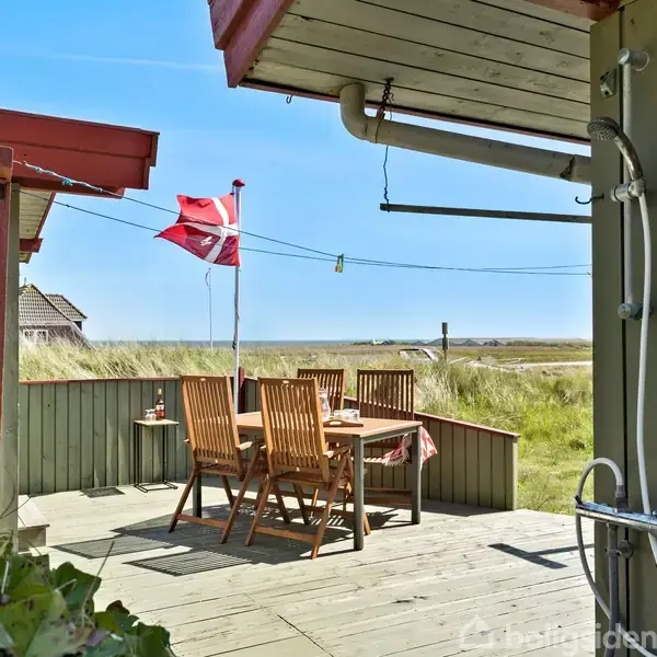 Udendørs træterrasse med bord og seks stole, udsigt over græsbevokset klitområde. Et dansk flag vajer fra en flagstang, og en udendørs bruser er synlig i højre side.