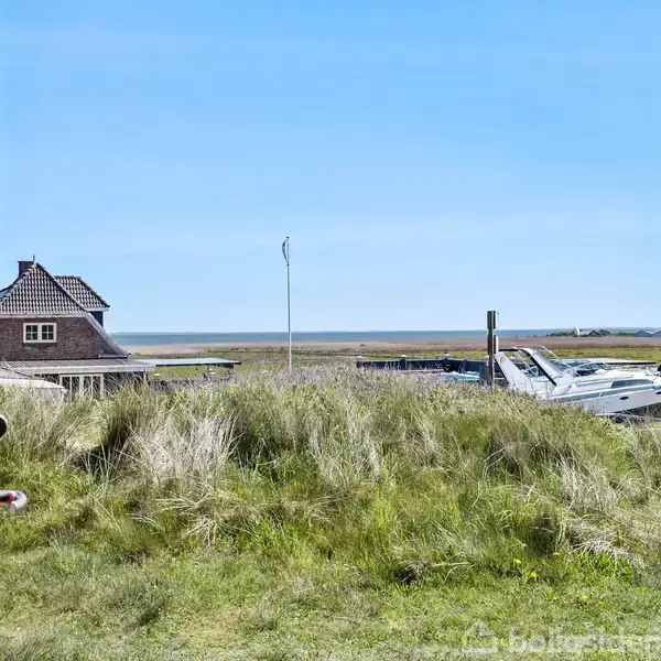 En rød dør står åben ud til en græsbevokset bakke med udsigt over et kystområde, hvor en båd og et hus med stråtag kan ses under en klar blå himmel.
