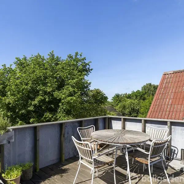 Rundt havebord med fire hvide stole står på en træterrasse. Terrasse omgivet af grønne træer og delvis udsigt til et rødt tegltag under en klar blå himmel.