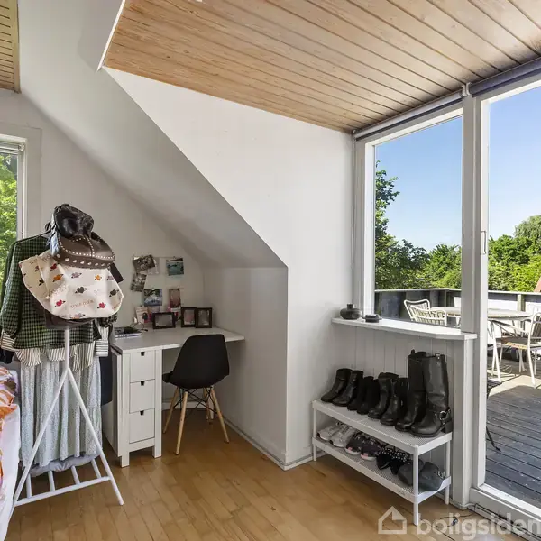 Soveværelse med seng til venstre og skrivebord ved vindue. Hatte hænger på stativ, sko er opstillet på reol. Terrasse udenfor med havemøbler, omgivet af træer og rødt tag.