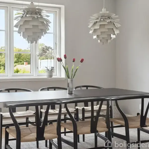 Spisebord med sorte stole står stille i et lyst rum med to hængende lamper. En vase med røde tulipaner pynter bordet, mens grønne træer ses gennem vinduet.