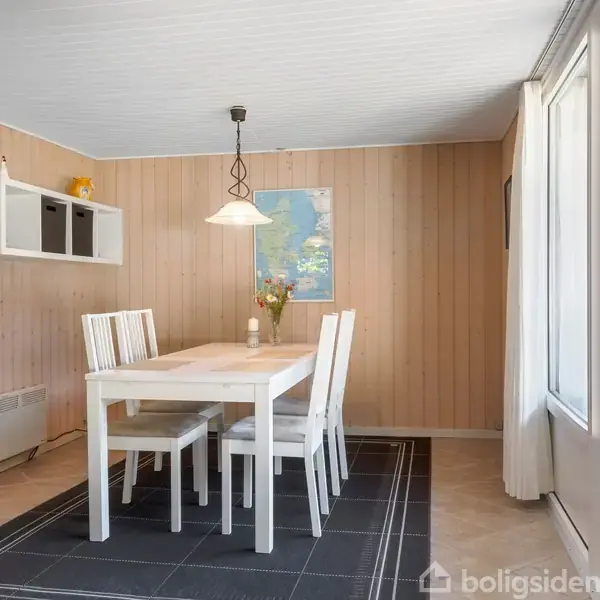 Et hvidt spisebord med fire stole står på et sort tæppe i et rum med trægulve; en lampe hænger over bordet. Væggen har træpaneler og er dekoreret med en plakat.