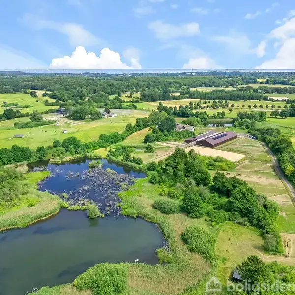 En stor sø omgivet af frodig grøn vegetation i et åbent landskab. I det fjerne ses landbrugsbygninger og marker, der strækker sig mod horisonten under en blå himmel.
