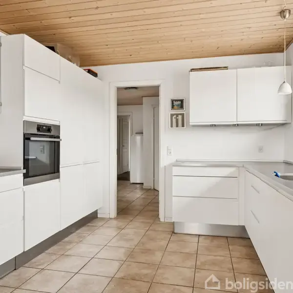 Hvidt køkken med moderne skabe, indbygget ovn og stålbordplade. Rummet har trægulv, loft i lyst træ og store vinduer. En blomsterpotte står ved vindueskarmen.