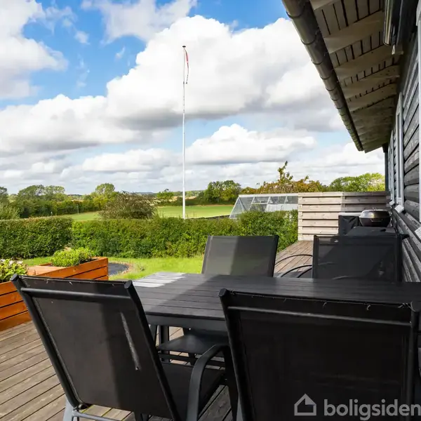Terrassebord med stole står på trædæk. Bordet er omkranset af grønt haveområde med flagstang og hæk. Himmel med skyer ses i baggrunden.