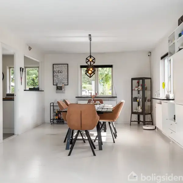 En spisestue med seks brune stole rundt om et spisebord. Der hænger en geometrisk lampe ovenover. Rummet har hvide vægge og vinduer, med dekorative billeder og hylder.