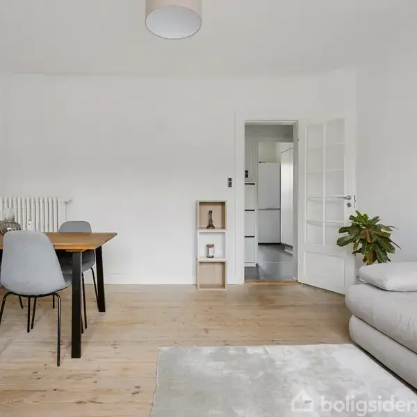 En stue med et lys træbord og fire grå stole ved siden af en lys sofa med puder. En plante står ved siden af døren, der fører til et andet rum.
