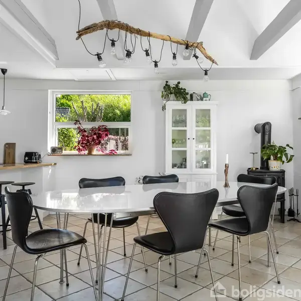 Et moderne køkken-alrum med sort-hvid indretning. Et ovalt spisebord med sorte stole står på flisegulvet. En stor plante og hvid skænk ses i baggrunden, omgivet af vinduer.
