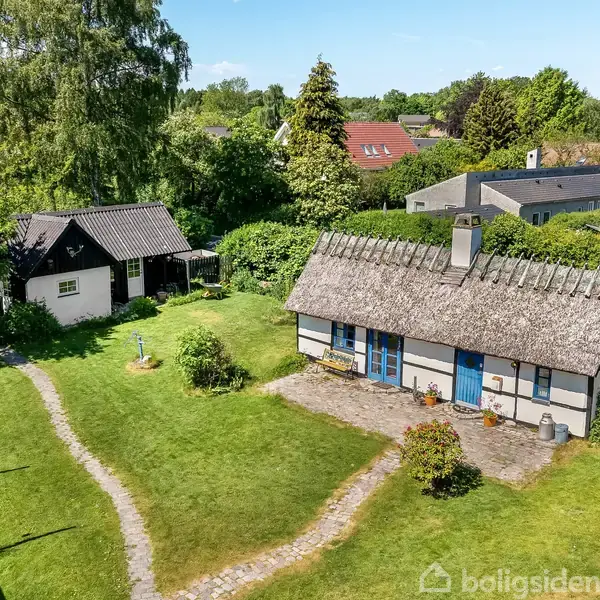 Hus med stråtag står på en grøn græsplæne med en fliseterrasse foran, omgivet af træer og buske. En sti fører til en mindre bygning med sort tag.