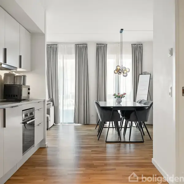 Køkken i moderne stil rummer hvide skabe og indbyggede apparater, som fører til en spisestue med et sort bord og stole. Tykke grå gardiner hænger i baggrunden. Vaskemaskine placeret til højre.