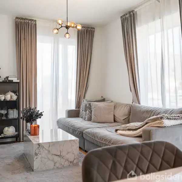Grå sofa med puder står i lys stue, omgivet af gardiner og glasvitrine med porcelæn. En marmor sofabord og orange vase med blomster placeret foran sofaen.