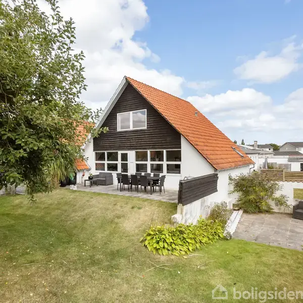 Hus med orange tegltag og hvid facade står i en have. Terrassen har havemøbler. Omkring er grønne træer og græsplæne. Skyer fylder himlen.