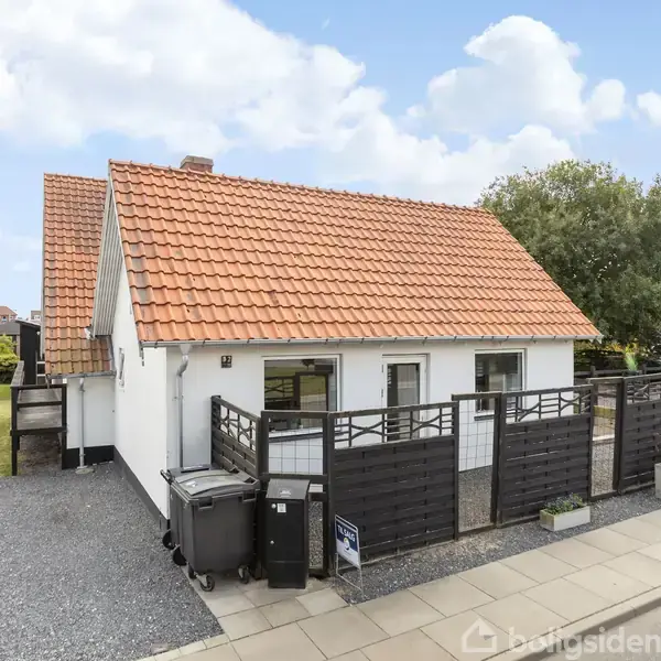 Huset har et rødt tegltag, hvide vægge og sorte vinduer, omgivet af et hegn med grusbelagt indkørsel. Det står i et beboelsesområde, med en græsplæne ved siden af.
