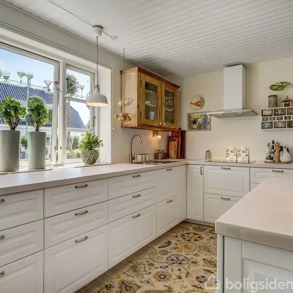 Hvidt køkken med store vinduer fyldt med planter og lys. Skabe langs væggene, kogeplade med emhætte, og ovn indbygget. Dekoreret med fliser på gulvet og belysning fra loftet.