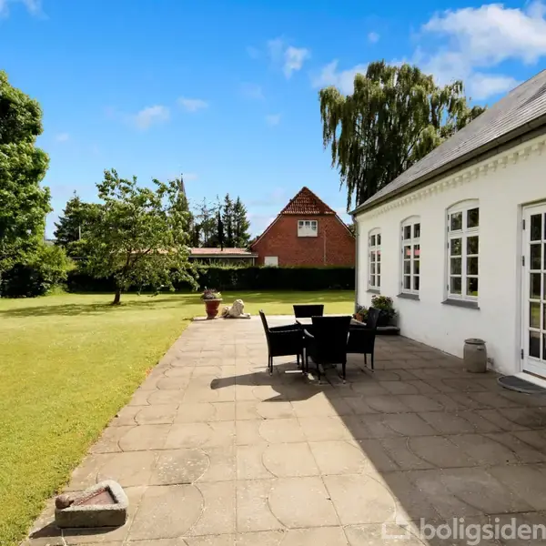 Hvid bygning med sorte tagplader, bord og stole på flisebelagt terrasse, grill tæt ved huset. Stor græsplæne med træer og en rød bygning i baggrunden under en blå himmel.