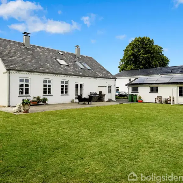 Hvidt hus med gråt tag står på en velplejet græsplæne. Til højre er en tilbygning med solpaneler og terrassemøbler. Blå himmel og træer i baggrunden.