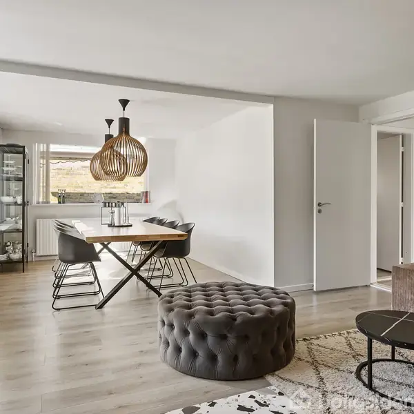 En rummelig stue med et rundt, mørkt puf foran et brunt sofaarrangement. I baggrunden står et spisebord med seks stole under to hængelamper i et lyst rum med store vinduer.