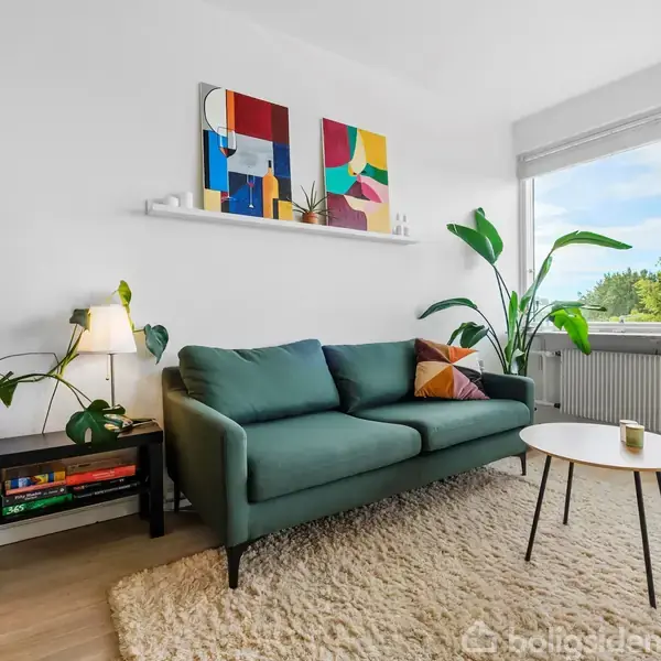 Grøn sofa står i lys stue med tæppe og sofabord, omgivet af planter. En hylde med farverige billeder hænger på væggen over sofaen. Vindue viser træer udenfor.