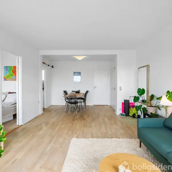 En stue med en grøn sofa og stuebord til højre, en fjernsyn ved en plante til venstre og spisestue med bord og stole i baggrunden. Lys, skandinavisk indretning.