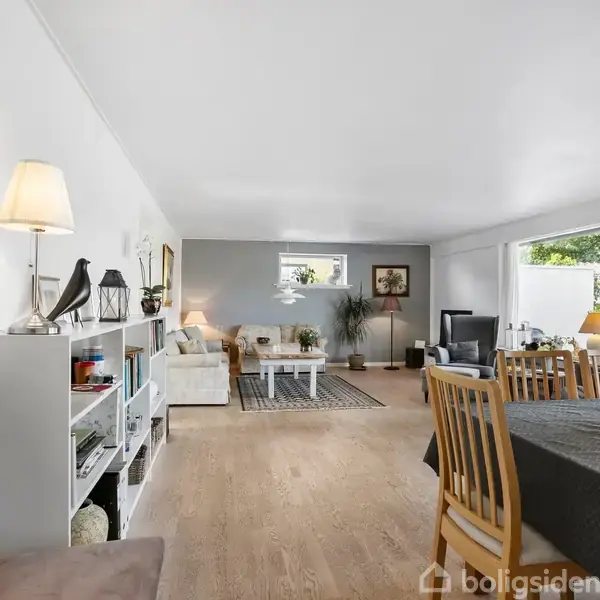 Stue med spisebord og stole står på trægulv, omgivet af hvide vægge og store vinduer. En sofa og reol med lamper og dekorationer supplerer rummet.