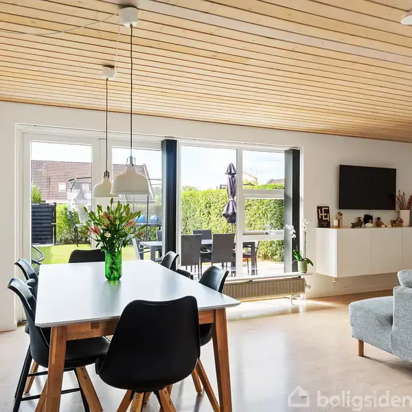 Et lyst spiseområde med et rektangulært bord omkranset af sorte stole. I baggrunden står en grå sofa og en terrasse med grønne omgivelser ses gennem store vinduer.