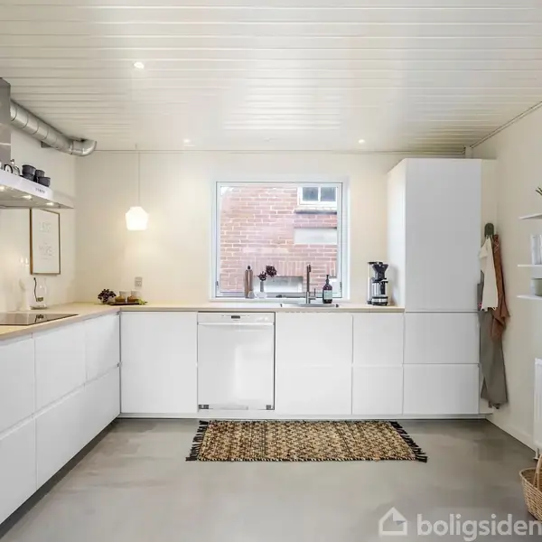 Lyst køkken med hvide skabe placeret i en U-form, aktion sker omkring et centralt vindue med udsigt til en murstensbygning. Konteksten inkluderer hylder, en ovn og en væglampe.