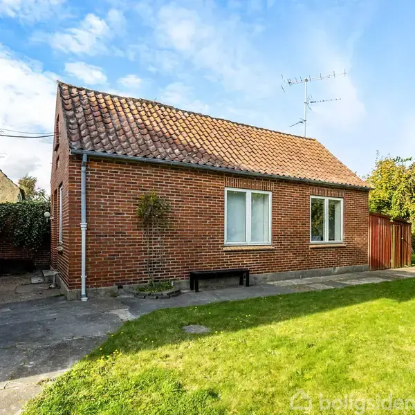 Et rødt murstenshus står stille i sollys. Det har et rødt tegltag, flankeret af en flisebelagt terrasse og en velplejet grøn græsplæne.