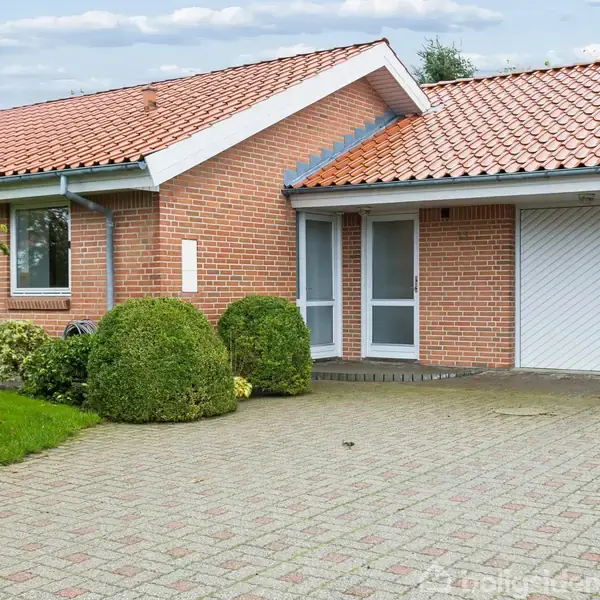 En rødmurstensvilla med rødt tegltag har en indkørsel af grå belægningssten. Bred hæk omkranser huset, og garagen ses til højre. klar, blå himmel i baggrunden.