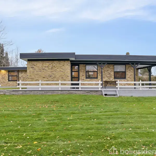 Et moderne étplanshus står midt på en velplejet græsplæne, omgivet af få træer. Huset har gul murstensfacade, sorte vinduer og en veranda med hvidt rækværk.