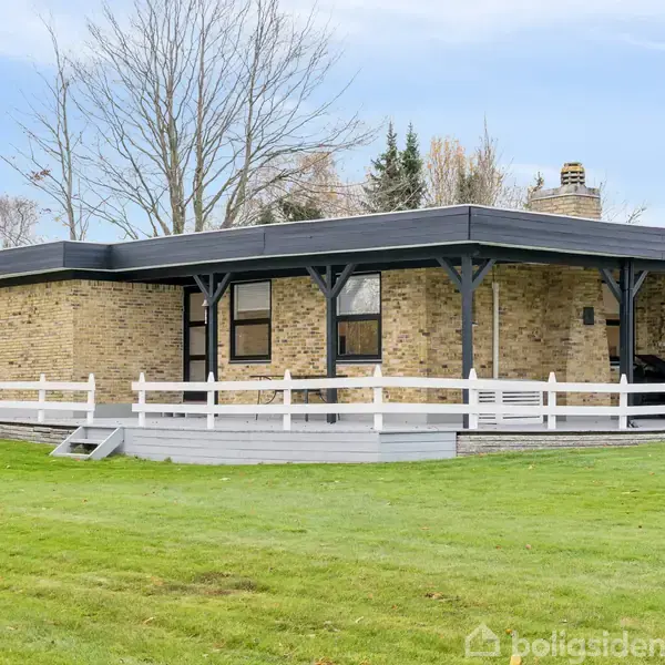 Et moderne hus står stille omgivet af en grøn græsplæne og nøgne træer. Bygget af lyse mursten med et fladt tag og en terrasse med hvidt rækværk.