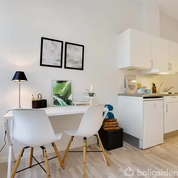To stole står ved et skrivebord med en lampe og dekorationer. I baggrunden er et lille køkken med skabe, vask og opbevaring. Lyse trægulve og hvidmalede vægge skaber et moderne look.