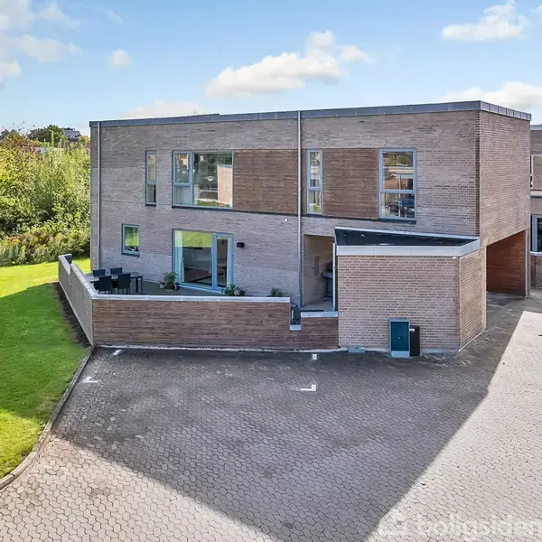 En moderne, tofamiliers villa i mursten med store vinduer står stille i et grønt boligområde. Huset har en flad tagkonstruktion og ligger ved en pæn stenlagt indgangsvej omgivet af græsplæner og træer.