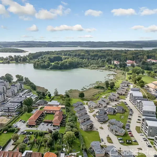 En gruppe boligbygninger ligger stille ved en sø omgivet af frodig skov. Bygningerne er moderne, mens søen og skoven skaber en naturskøn baggrund.