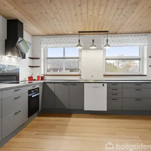Køkken med grå skabe og moderne apparater, herunder en indbygget ovn. Træloft og gulv skaber en varm atmosfære. To store vinduer slipper naturligt lys ind, omgivet af hylder.