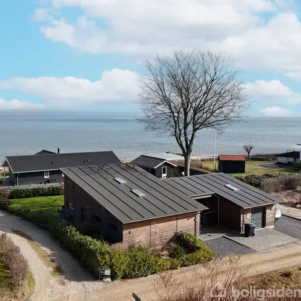 Et træhus står stille ved kysten, omgivet af flere småbygninger og en enkelt træ i en landsby nær havet. Skydækket himmel ses i baggrunden.