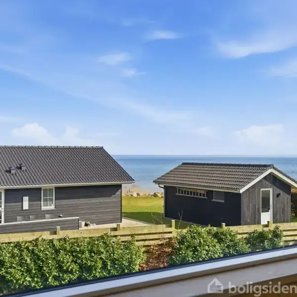 Et vindue indrammer udsigten til to sorte træhuse med græstag, der står i en have med havet i baggrunden under en blå himmel.