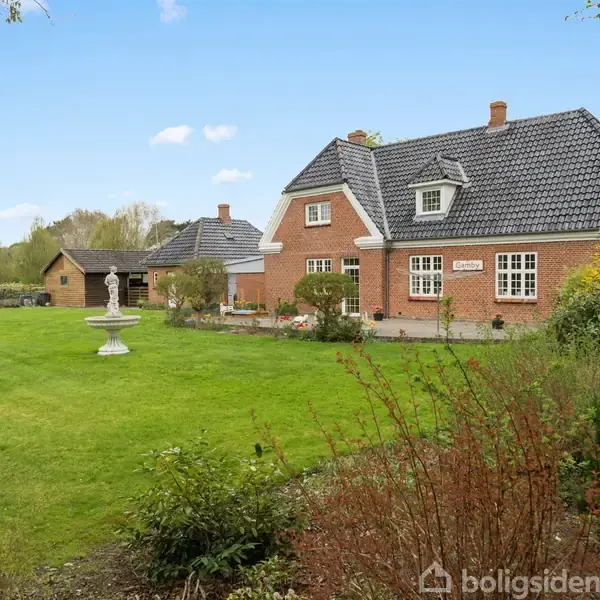 Et murstenshus med mørkt tag står i en velplejet have med græsplæne og en statue. Baggrunden viser træer og en lys, blå himmel.