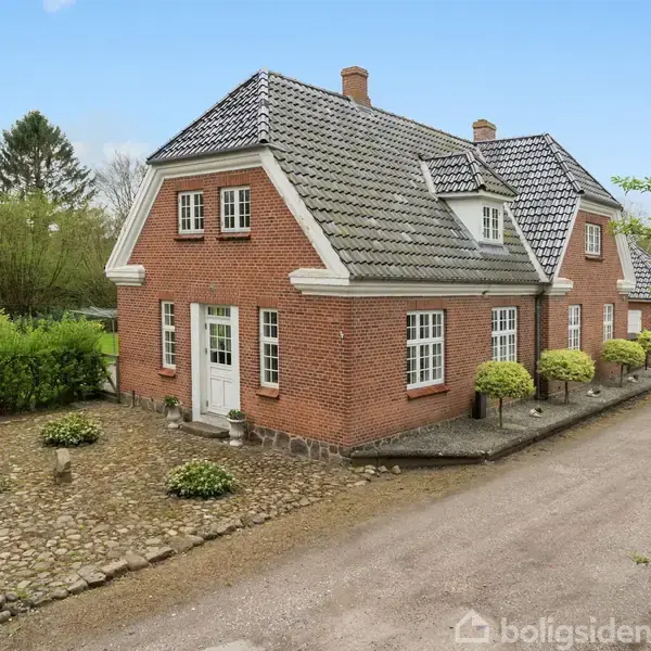 Rødt murstenshus med sort- og gråt tegltag står stille ved en grusvej. Omgivet af en have med buske og træer i en landlig, forårspræget setting.