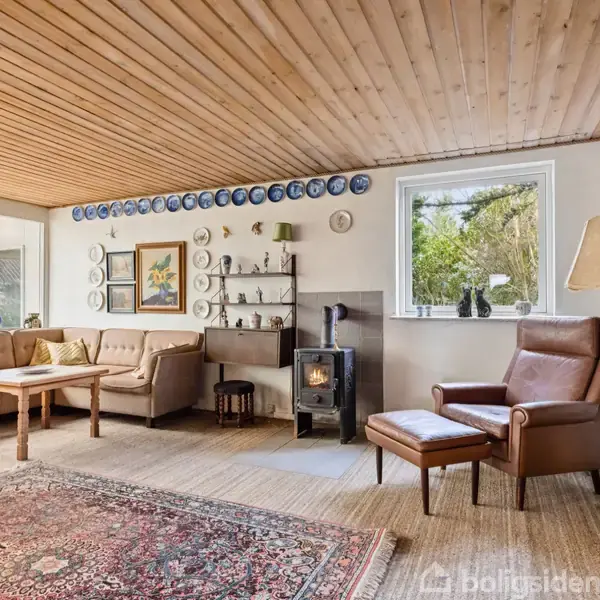Stue med brændeovn og lænestol ved siden af et vindue. Langs væggen står en sofagruppe med sofabord. Reoler med bøger til højre og tallerkener hænger over sofaen.